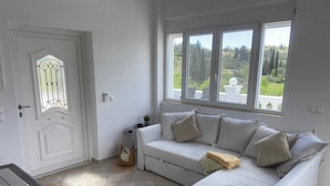 A white room with a white sofa, a door with a glass panel, and a window with a view of greenery. - Private Villa and Pool With Lake Views and Sunset Terraces at The Grove (Corfu)