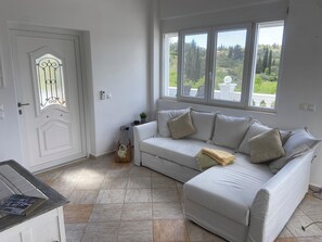 Ett vitt rum med en vit soffa, en dörr med ett glasparti och ett fönster med utsikt över grönområden.