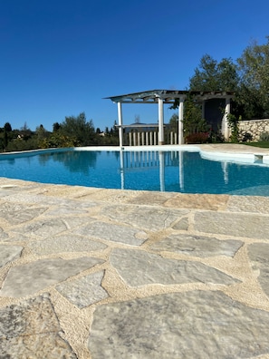 Ein Schwimmbecken mit Steinfliesen, einer Pergola und umgebendem Grün.