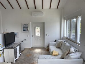 A living room with a white sofa, a television on a cabinet, a door with a glass panel, and a window with a view.