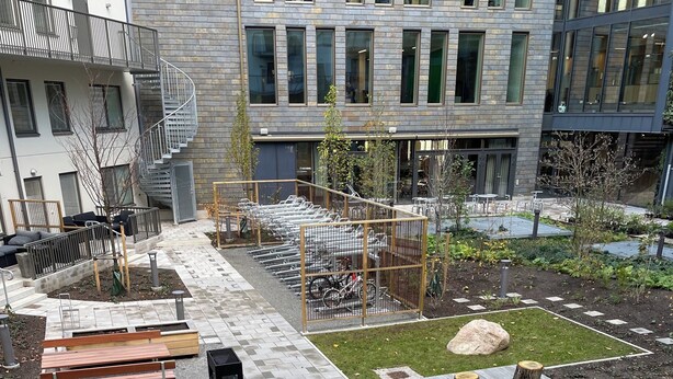 A courtyard with a paved walkway, benches, a small garden area with a sandbox, and a building with multiple windows and a staircase. - Hyllie Appartement - New Construction (Malmö)