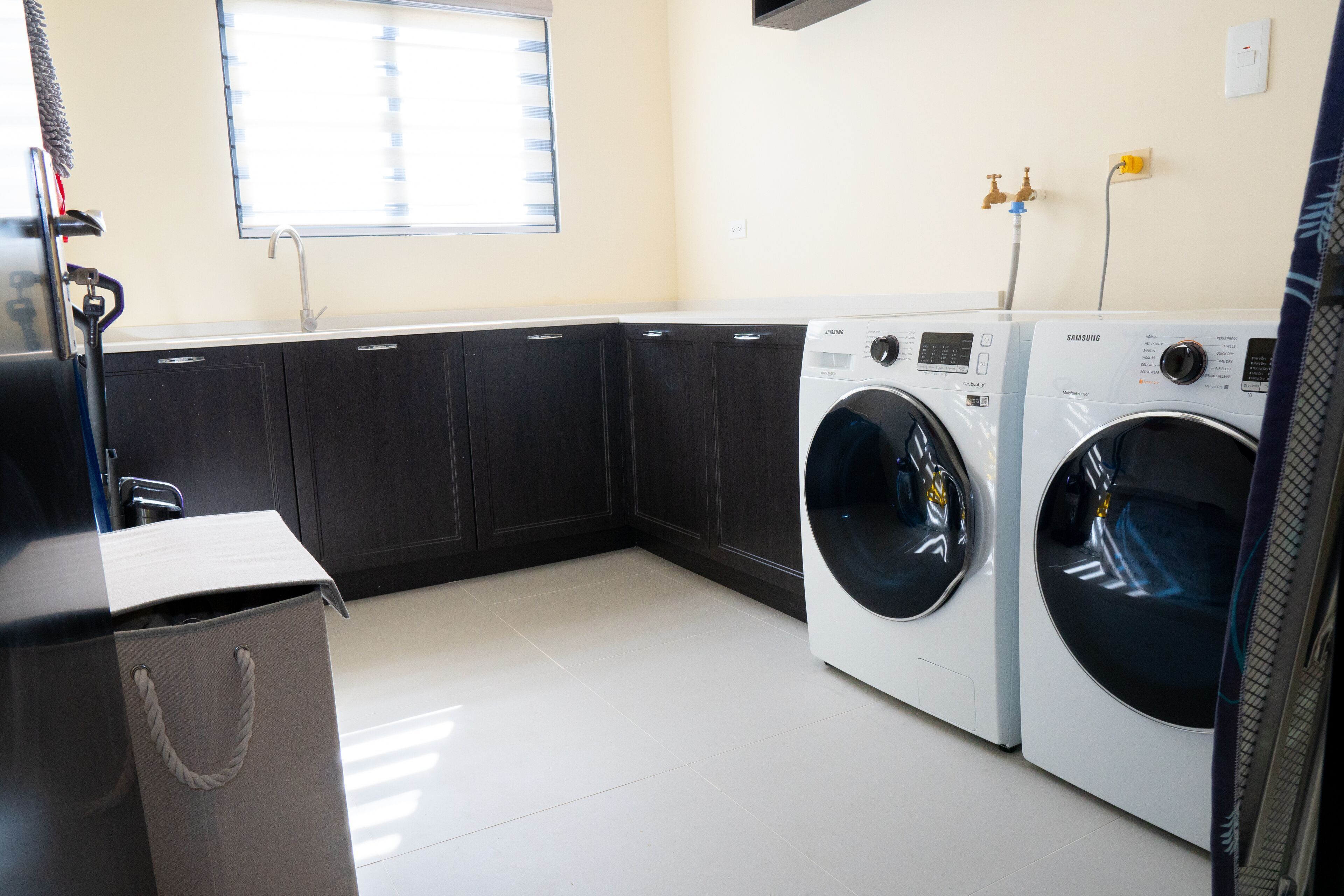 A laundry room with two white front-loading washers, a sink, and a window with blinds.