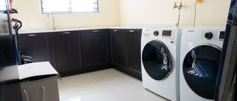 A laundry room with two white front-loading washers, a sink, and a window with blinds.