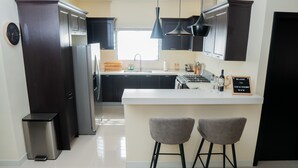 A modern kitchen with dark cabinetry, stainless steel appliances, a white countertop, and two bar stools. - Chic Hideaway a Paradise Home in Jamaica (Ocho Rios)