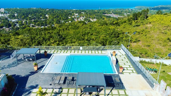 Une piscine sur le toit offrant une vue sur l’océan, entourée de verdure et d’une clôture blanche.