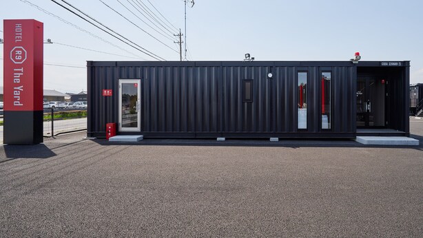Ein modernes, eingeschossiges Gebäude mit dunkler Fassade, einem Glastür-Eingang und einem sichtbaren Brandmelder.