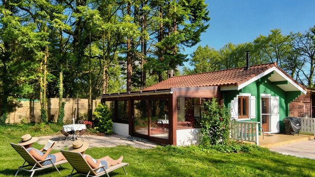 Una casa verde con techo de tejas, un porche con paredes de cristal y un césped con dos tumbonas.
