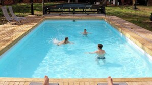 Una piscina con dos sillas reclinables en el borde, una persona nadando y otra leyendo un libro.