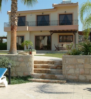 A two-story house with a stone staircase leading to the entrance, a balcony with a railing, and a patio area with outdoor furniture. - Three Bedroom Villa With Private Pool and Landscaped Garden Close to the Beach (Pissouri)