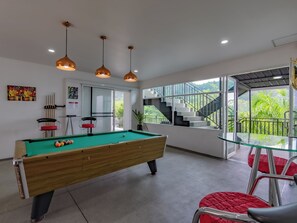 A pool table in a room with a glass table and red chairs, a staircase leading to a balcony with a view of greenery.