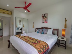 A hotel room with a bed, a ceiling fan, a bedside table with a lamp, a mirror, and a door leading to another room. - Villa Nap Dau Crown Private Pool Villa Phuket (Chalong)