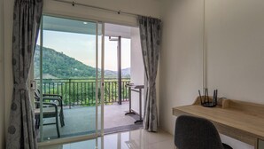 Ein Zimmer mit einem Holztisch, einem Stuhl, einem Balkon mit Blick auf Grünflächen und Hügel sowie einer Schiebetür.