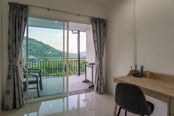 Habitació amb escriptori de fusta, cadira, balcó i vista a zones verdes i turons, i porta corredissa de vidre.