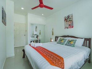 Een net opgemaakt bed met een rode decoratieve loper, een plafondventilator, een bloemschilderij en een spiegel aan de muur.