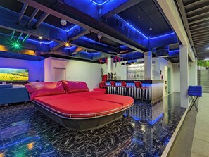 Une chambre d’hôtel moderne avec un lit rouge, un coin bar et un grand téléviseur diffusant un paysage pittoresque.