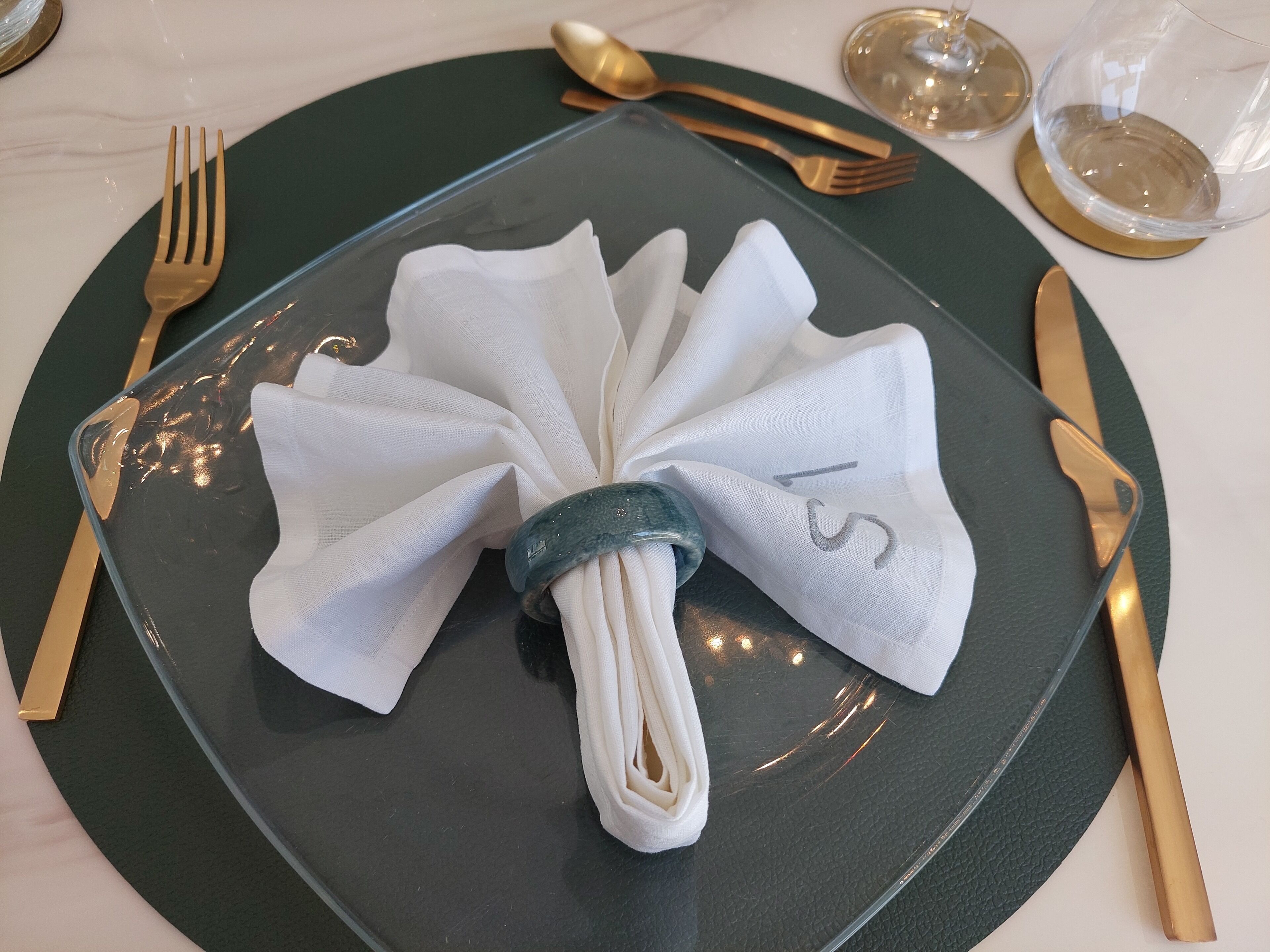 A neatly folded white napkin on a dark green plate, accompanied by gold cutlery and a glass of water on a white tablecloth.