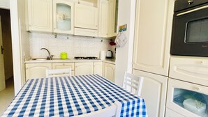 Una cocina con armarios blancos, una estufa y un lavamanos. Se observa una mesa con mantel a cuadros y dos sillas.