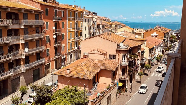 Filas de edificios con techos de teja anaranjada y balcones, una calle en primer plano y una vista despejada del mar al fondo.