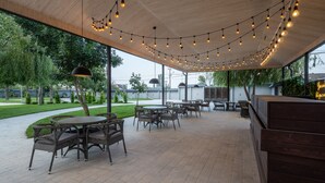 Un espace extérieur aménagé en terrasse, avec des tables rondes et des chaises en osier, des guirlandes lumineuses suspendues et une vue sur la verdure environnante.