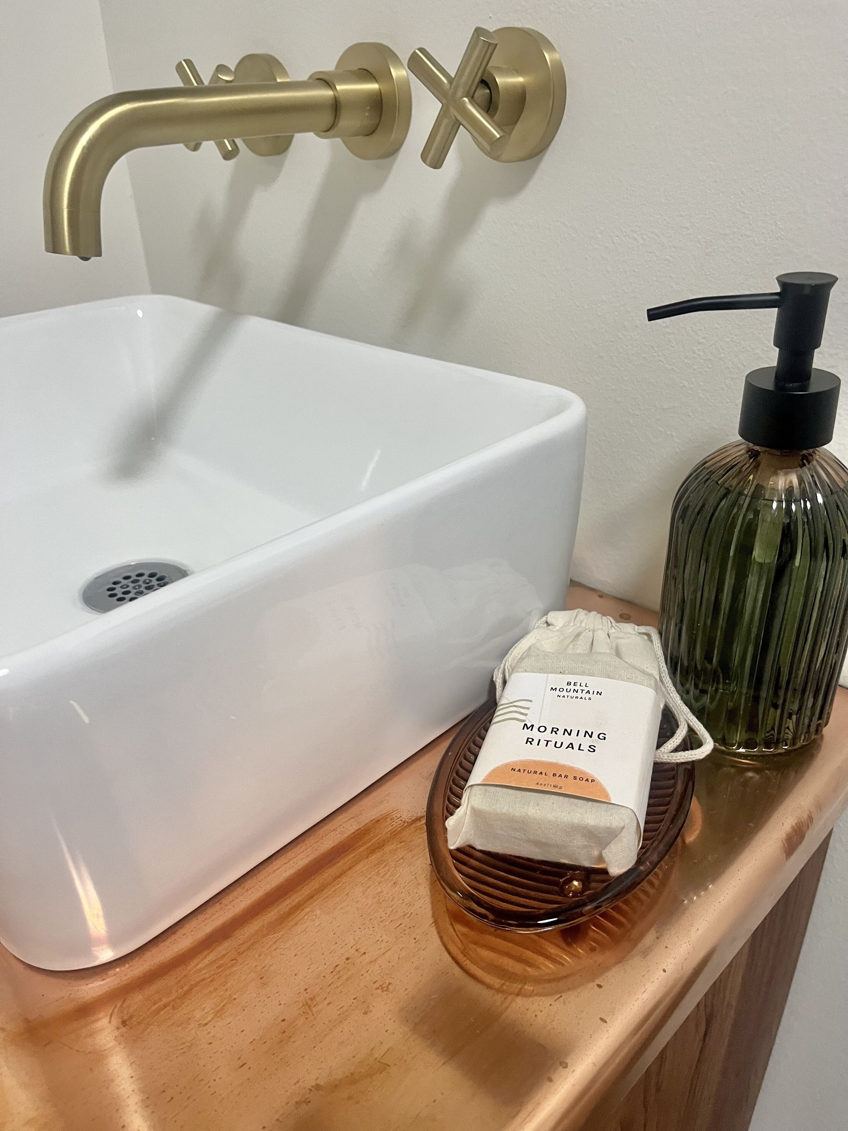 Un lavabo con un rubinetto dorato, un dispenser di sapone e un contenitore di vetro su un piano in legno.