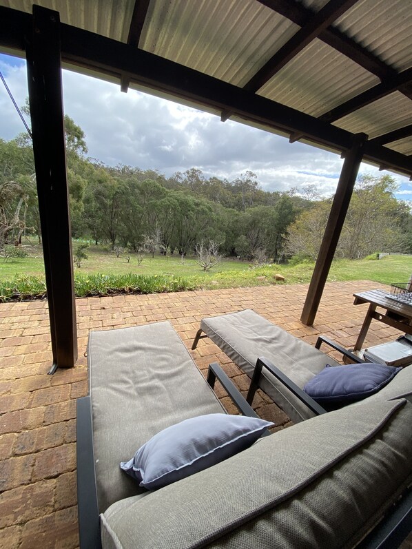 A covered outdoor seating area with two lounge chairs and a table, overlooking a grassy landscape and trees.