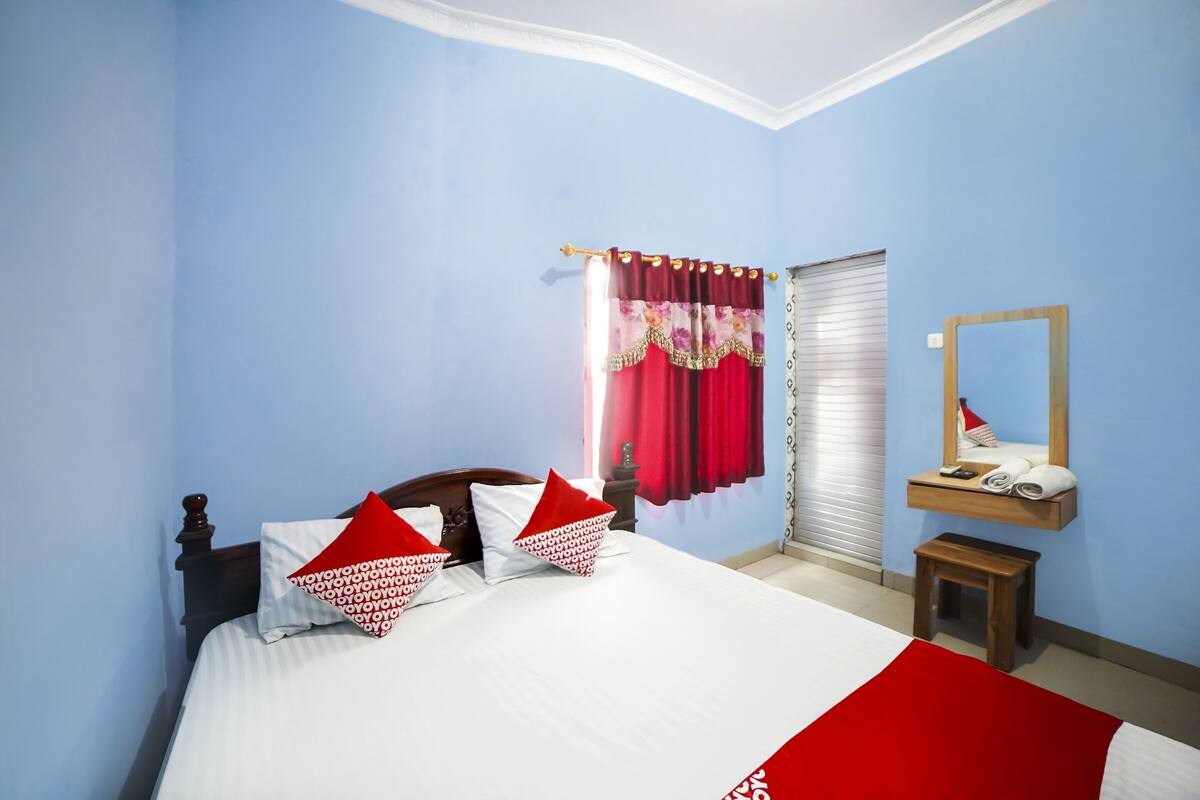 Quarto com cama, almofadas, mesa de cabeceira, espelho e janela com cortinas.