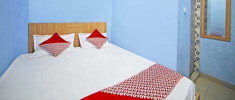 A hotel room with a bed, two pillows, a red and white patterned blanket, a wooden headboard, a wall-mounted air conditioner, and a mirror with a patterned frame.