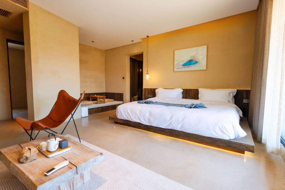 A hotel room with a large bed, a wooden coffee table with a remote control, a chair, and a small table with a teapot and cups.