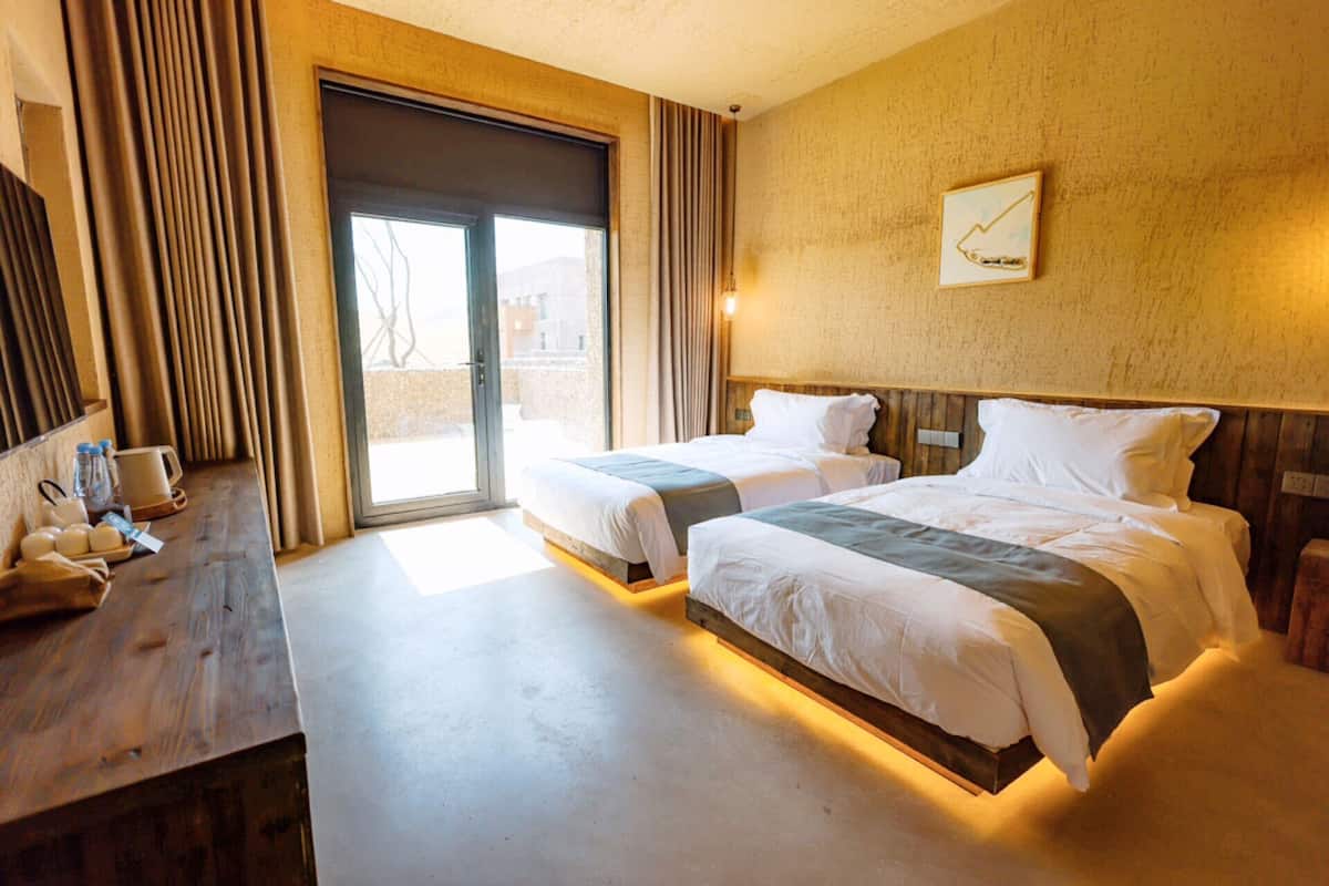 A hotel room with two beds, a wooden desk with amenities, a large window, and a wall-mounted artwork.