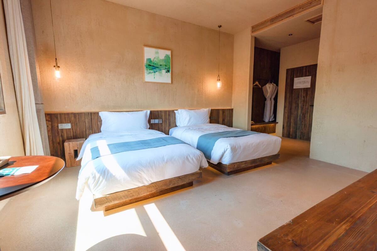 A hotel room with two beds, wooden headboards, a painting on the wall, and a wooden bench.