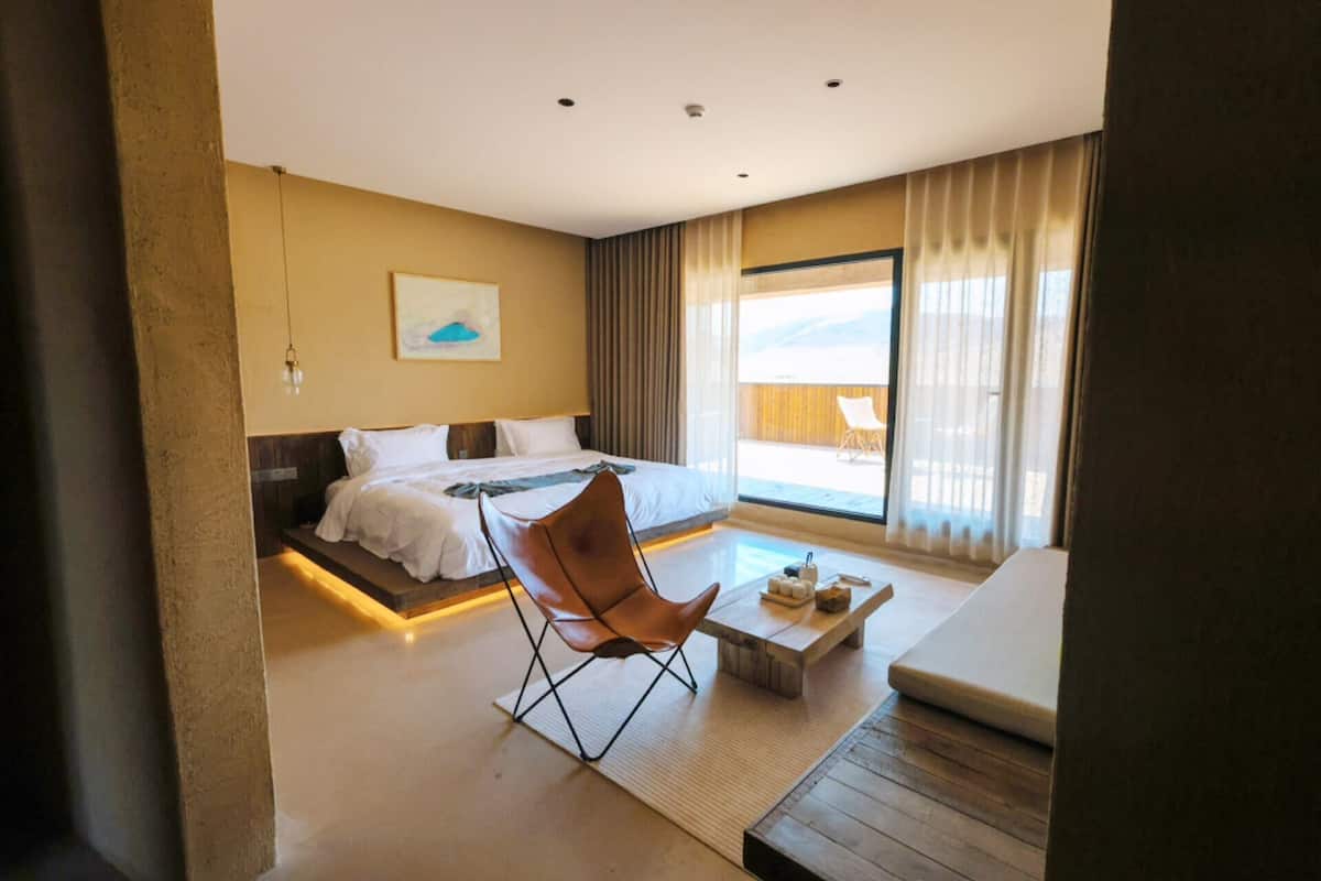 A modern hotel room with a bed, a seating area with a chair and table, and a large window with a view.
