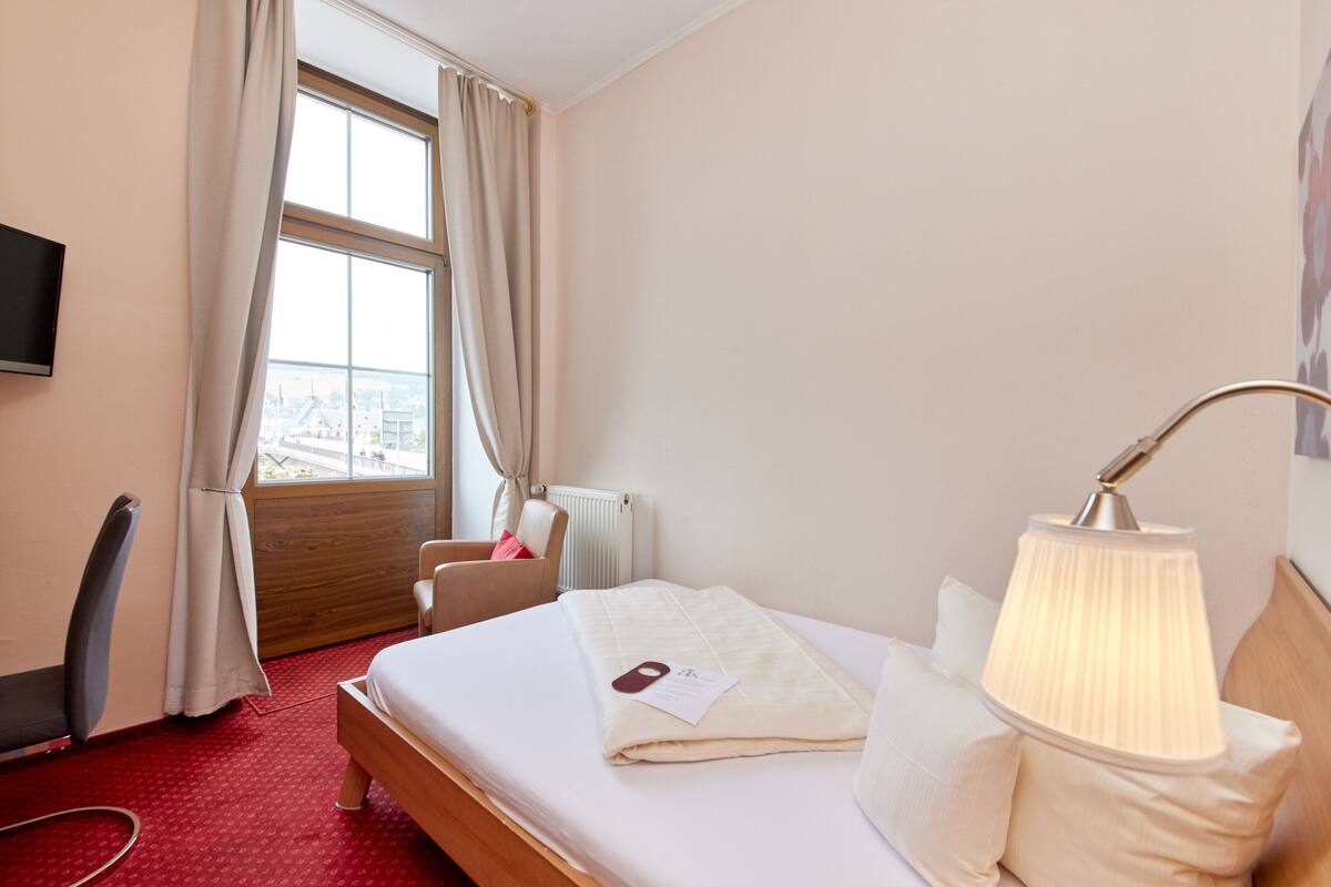 A hotel room with a bed, a desk, a chair, a lamp, and a window with curtains.