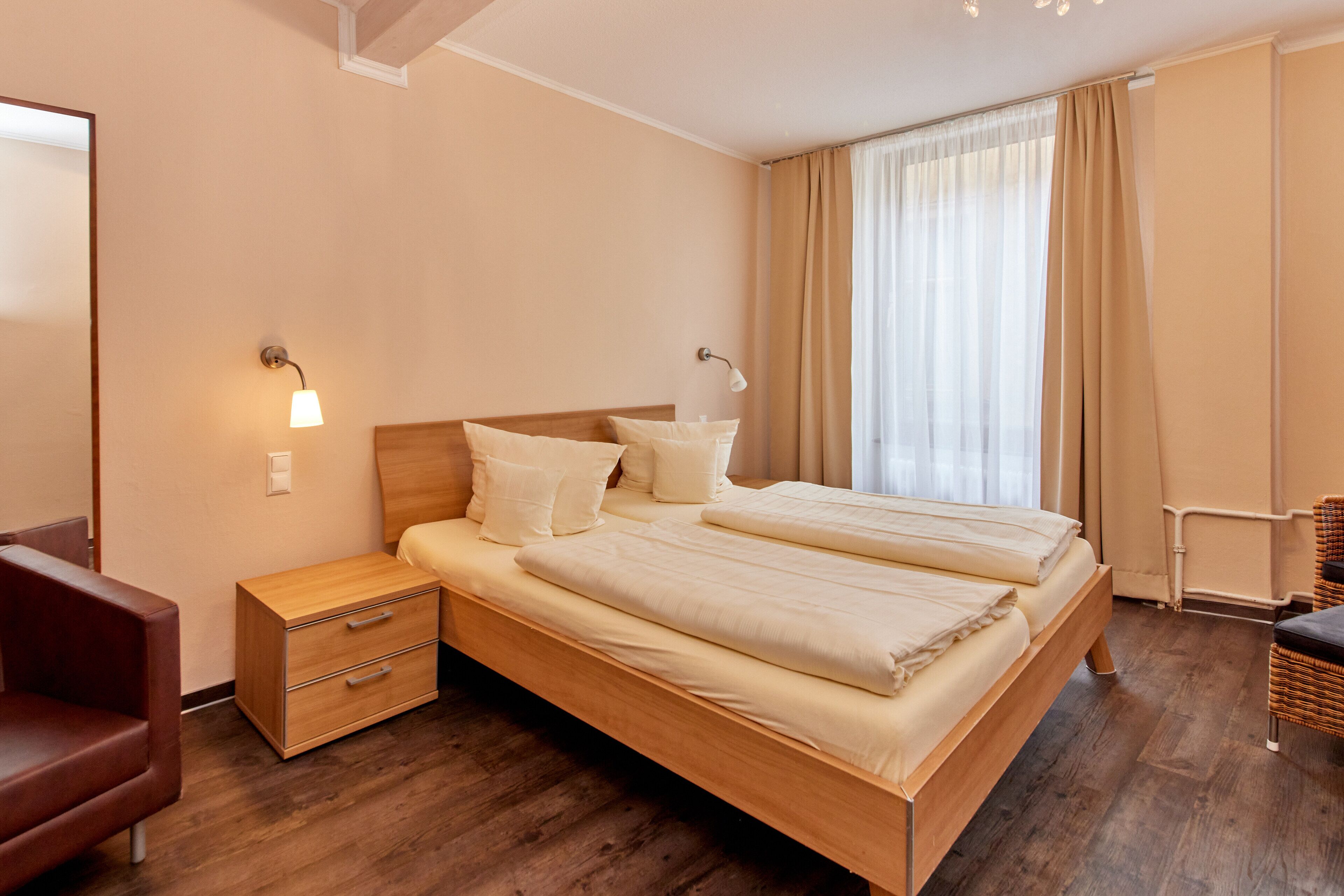 Una habitación de hotel moderna con una cama de madera, lámparas de noche, una mesita de noche, una silla y un ventanal con cortinas.