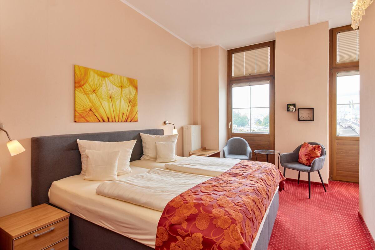 A hotel room with a large bed, two bedside tables, a chair, and a window with blinds.