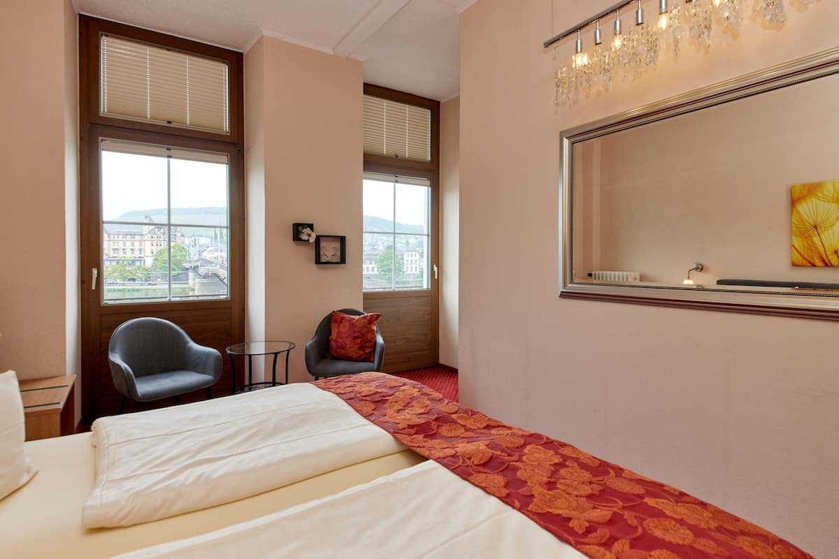 A hotel room with a bed, two chairs, a small table, and a large window with blinds.