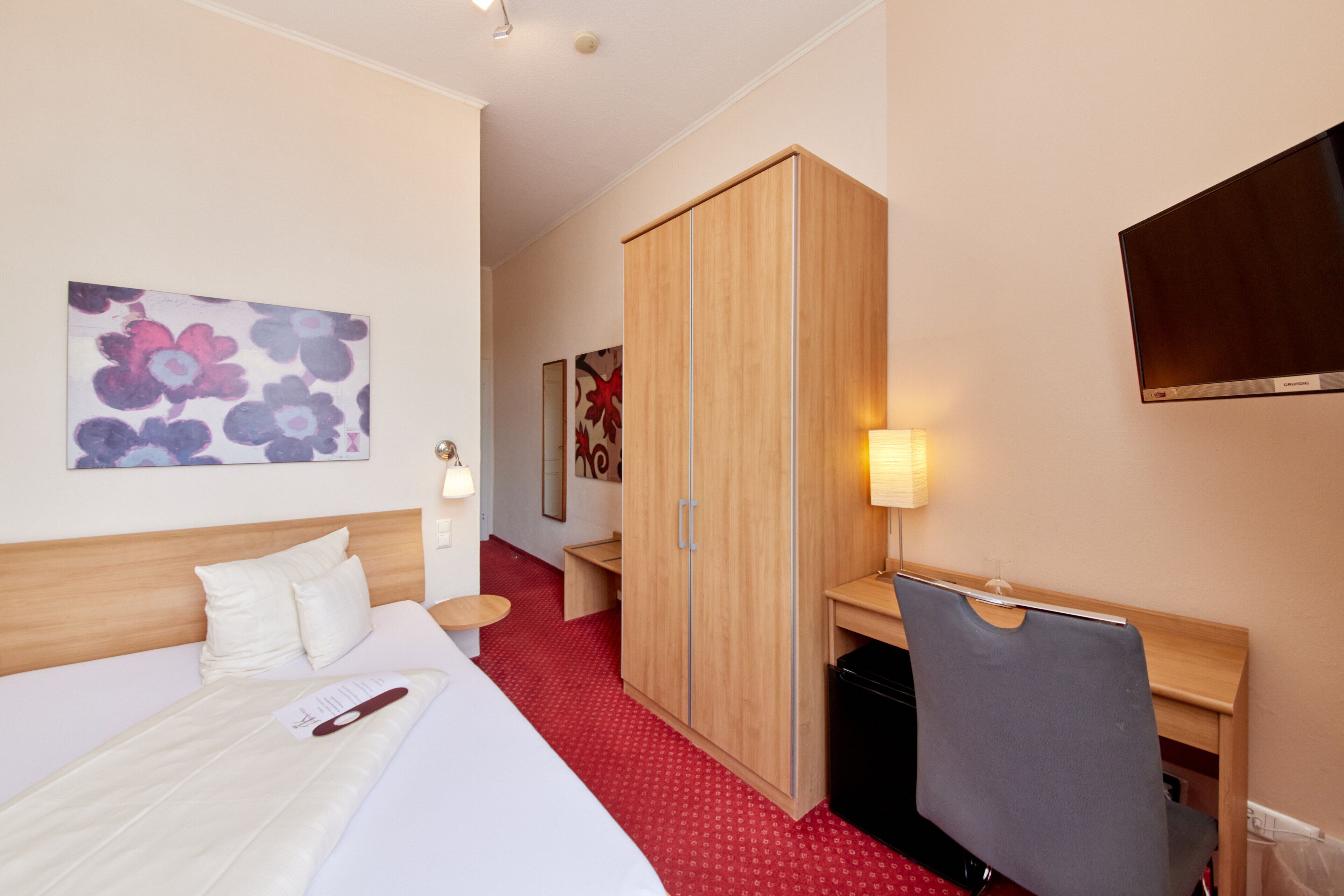 A hotel room with a bed, a desk, a wardrobe, a TV, and artwork on the walls.