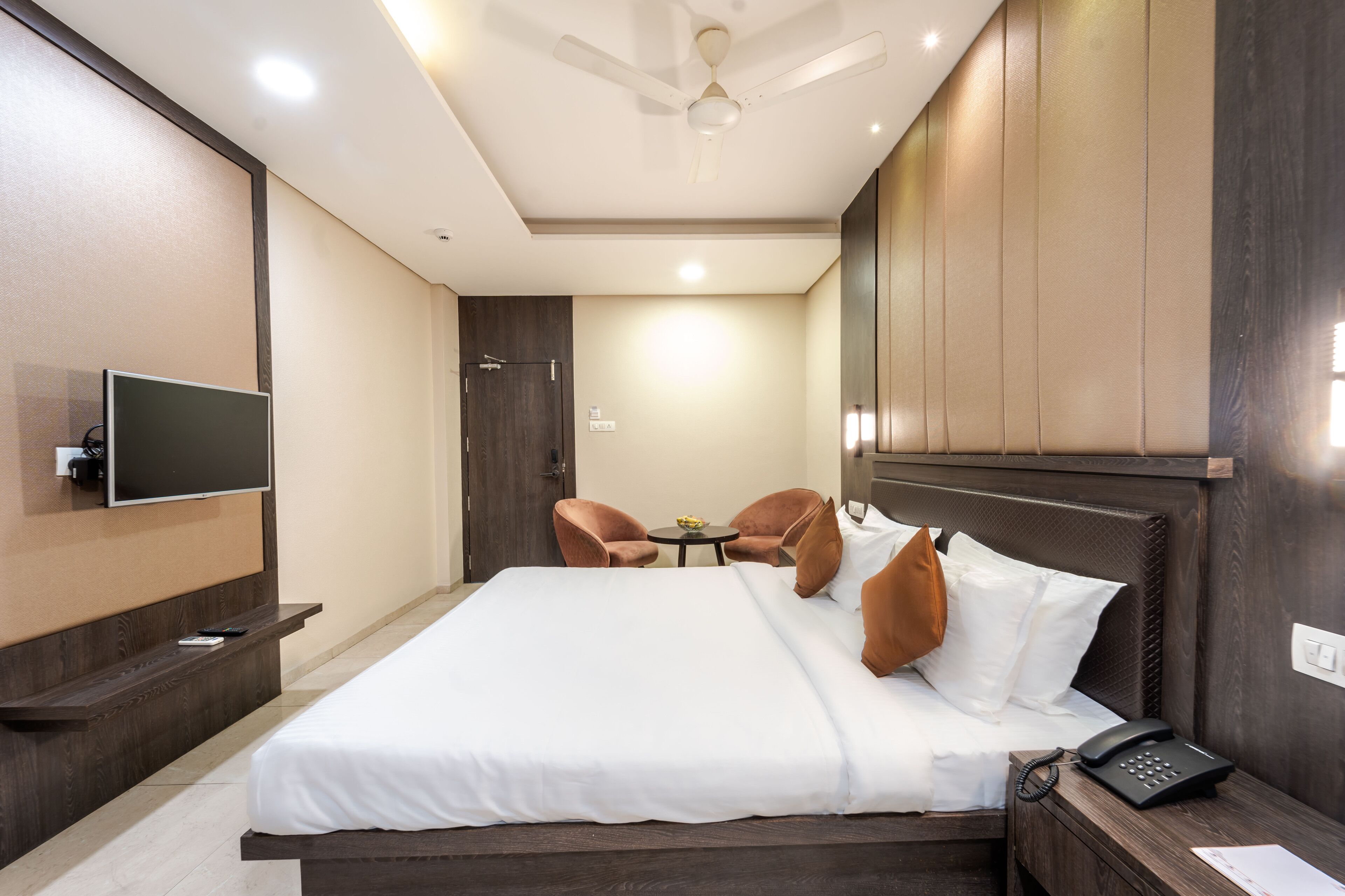 Una habitación de hotel con una cama grande, un televisor montado en la pared, un escritorio con teléfono y un ventilador de techo.