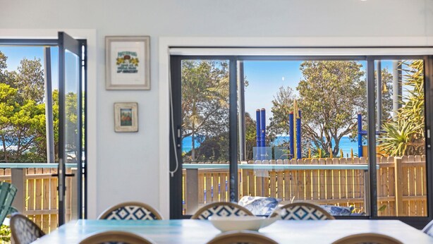 Uma área de refeições com uma mesa grande e cadeiras, janelas amplas com vista para árvores e um cercado, e quadros nas paredes.