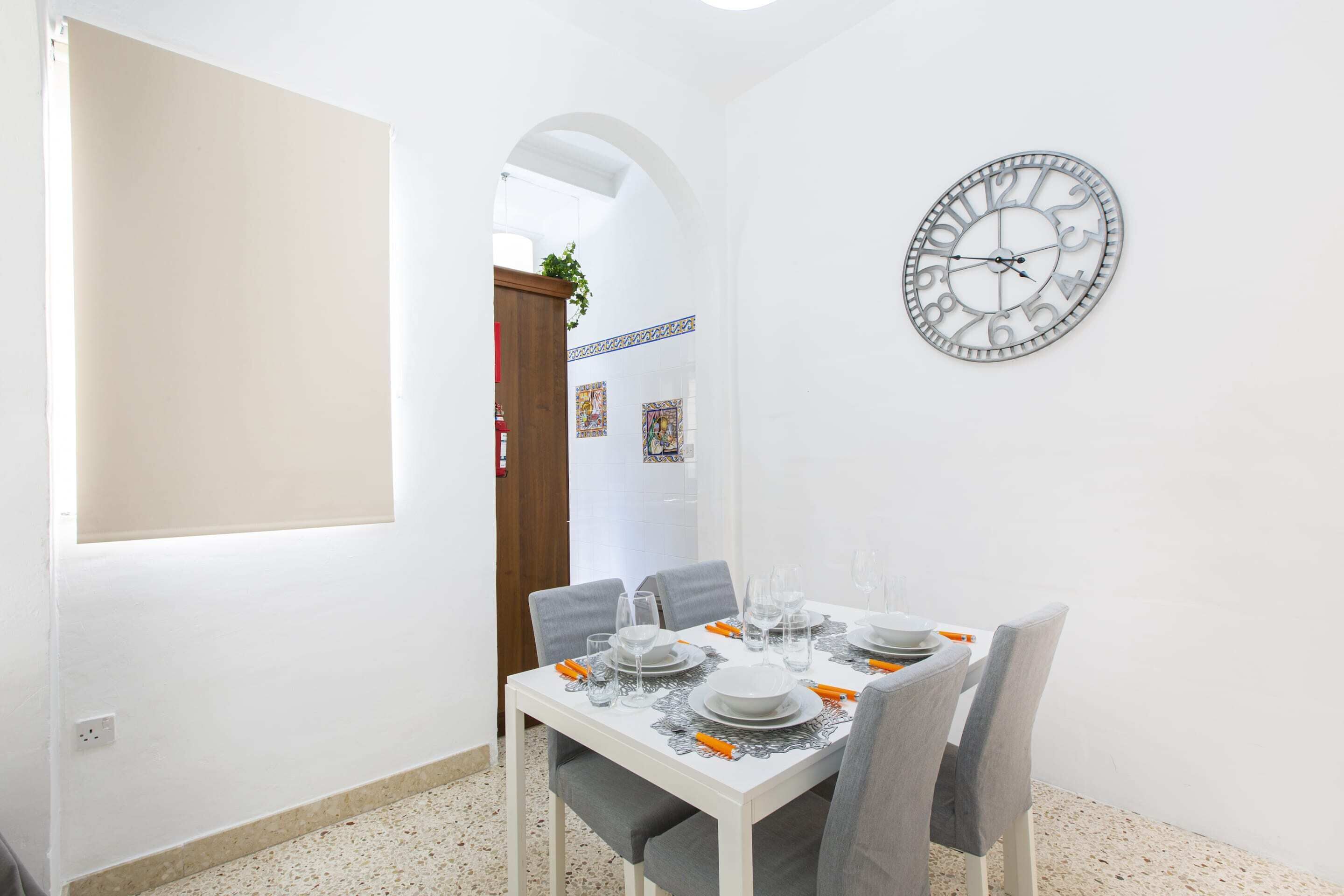 A dining area with a white table set for four, a wall clock, and a door leading to another room.
