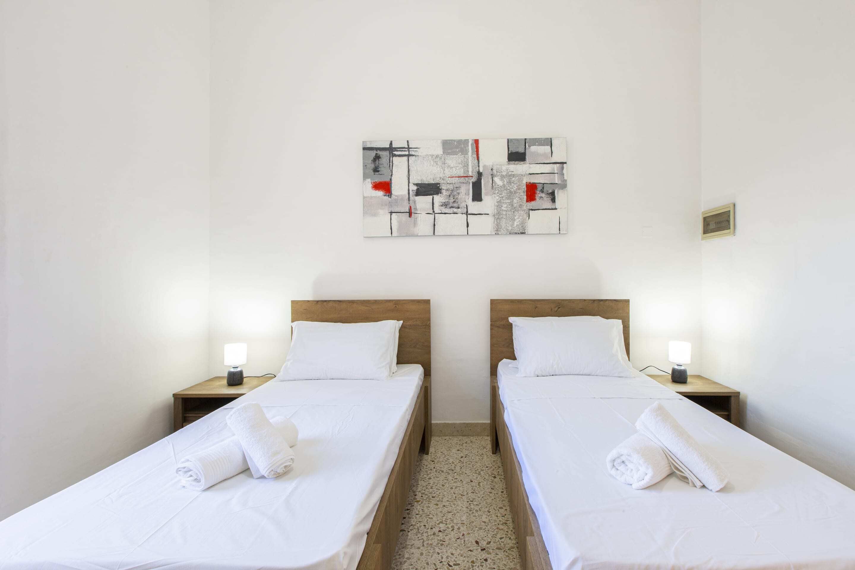 Habitación de hotel con dos camas individuales, cada una con cabecero de madera, ropa blanca y una toalla enrollada sobre la cama. Una mesita pequeña se encuentra entre las camas con una lámpara encima. Sobre las camas, un cuadro abstracto enmarcados decora la pared.