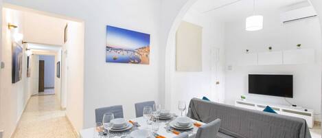 Un comedor moderno con una mesa preparada para cuatro personas, un sofá y un televisor al fondo.