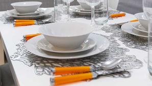 A dining table set with white plates, bowls, and clear wine glasses, with orange handles on the cutlery. - Sliema Lovely Flat (Sliema)