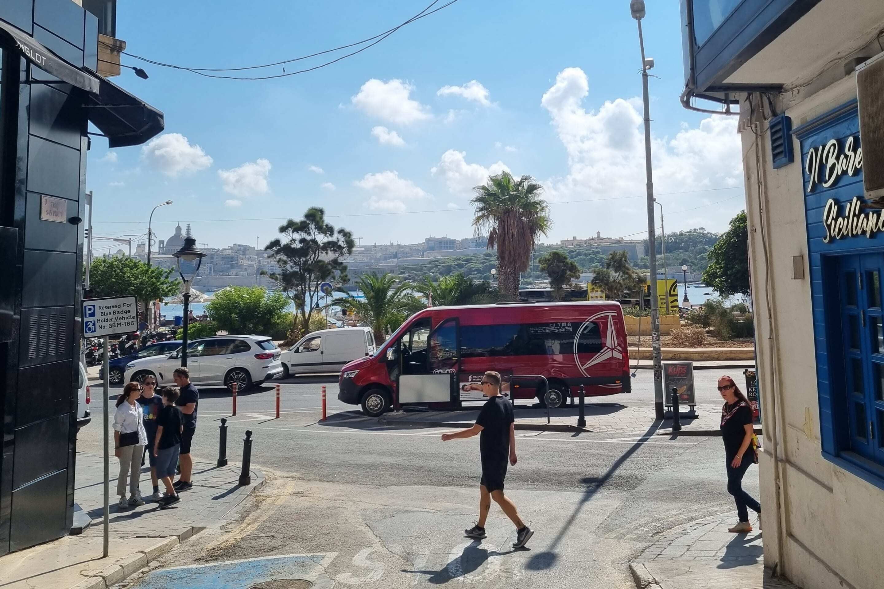 A street scene with a red van parked on the side, a blue building with a sign, and a few pedestrians.