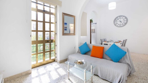 A living room with a sofa, a glass coffee table, a wall clock, and a door leading to an outdoor area. - Sliema Lovely Flat (Sliema)