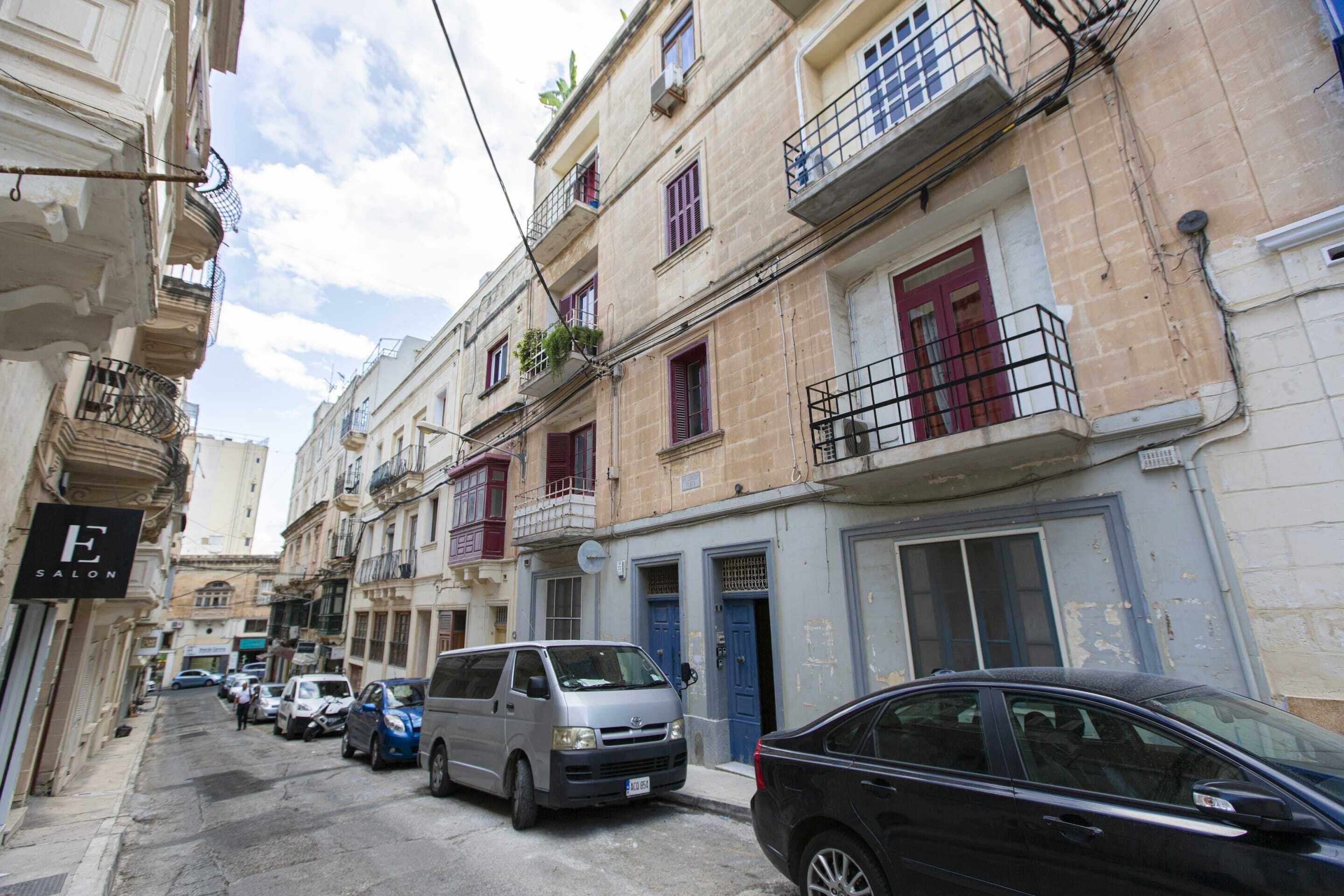 A narrow street lined with multi-story buildings, some with balconies and others with ground-level shops.
