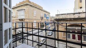 A balcony with a view of a street, buildings, and a car parked on the street. - Sliema Lovely Flat (Sliema)