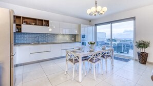 A modern kitchen with white cabinets, a blue-tiled backsplash, and a dining area with a white table and chairs. - Pool Penthouse (Swieqi)