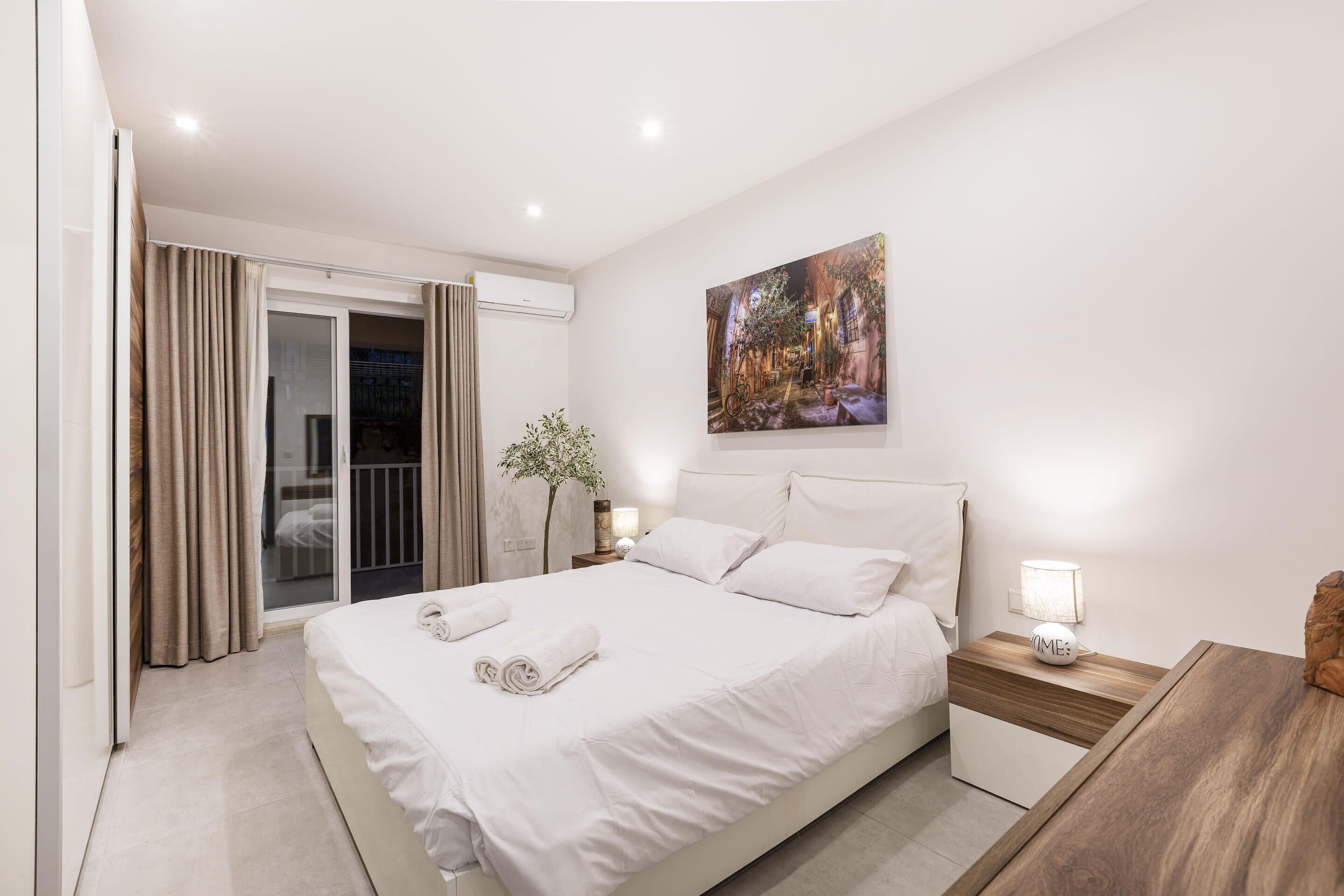Una habitación de hotel moderna con una cama grande, una mesita de noche con lámpara, un cuadro en la pared y vistas a través de la puerta de cristal.