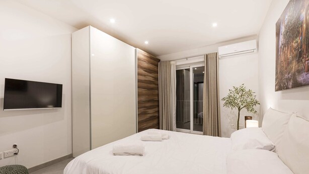 A modern hotel room with a large bed, a flat-screen TV mounted on the wall, a sliding door to a balcony, and a potted plant by the window. - Golden Flat in San Pawl (St. Paul's Bay)