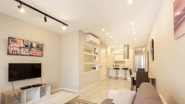 A modern living room with a flat-screen TV, a wooden coffee table, and a brown sofa. There is a kitchen area in the background with a dining table and chairs. - Brand new 1BR in Central Malta (Pieta)